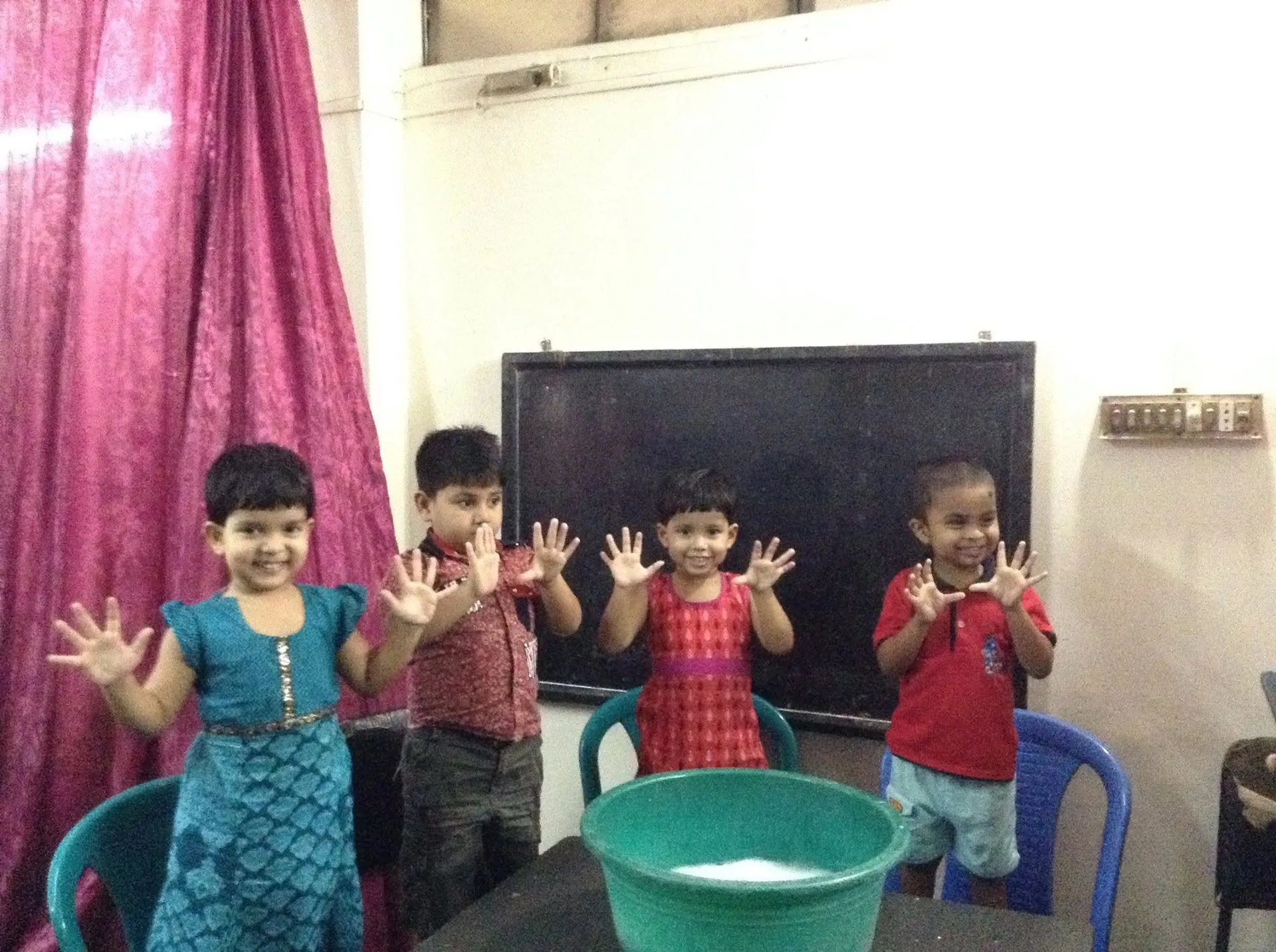 Four smiling young children hold up their clean hands beside a basin of water in a classroom
