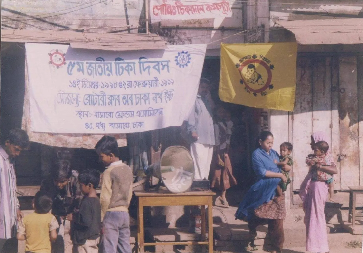 Mothers and children at a street-side booth under a banner for the fifth National Immunization Day, held at BFA, in an old printed photograph