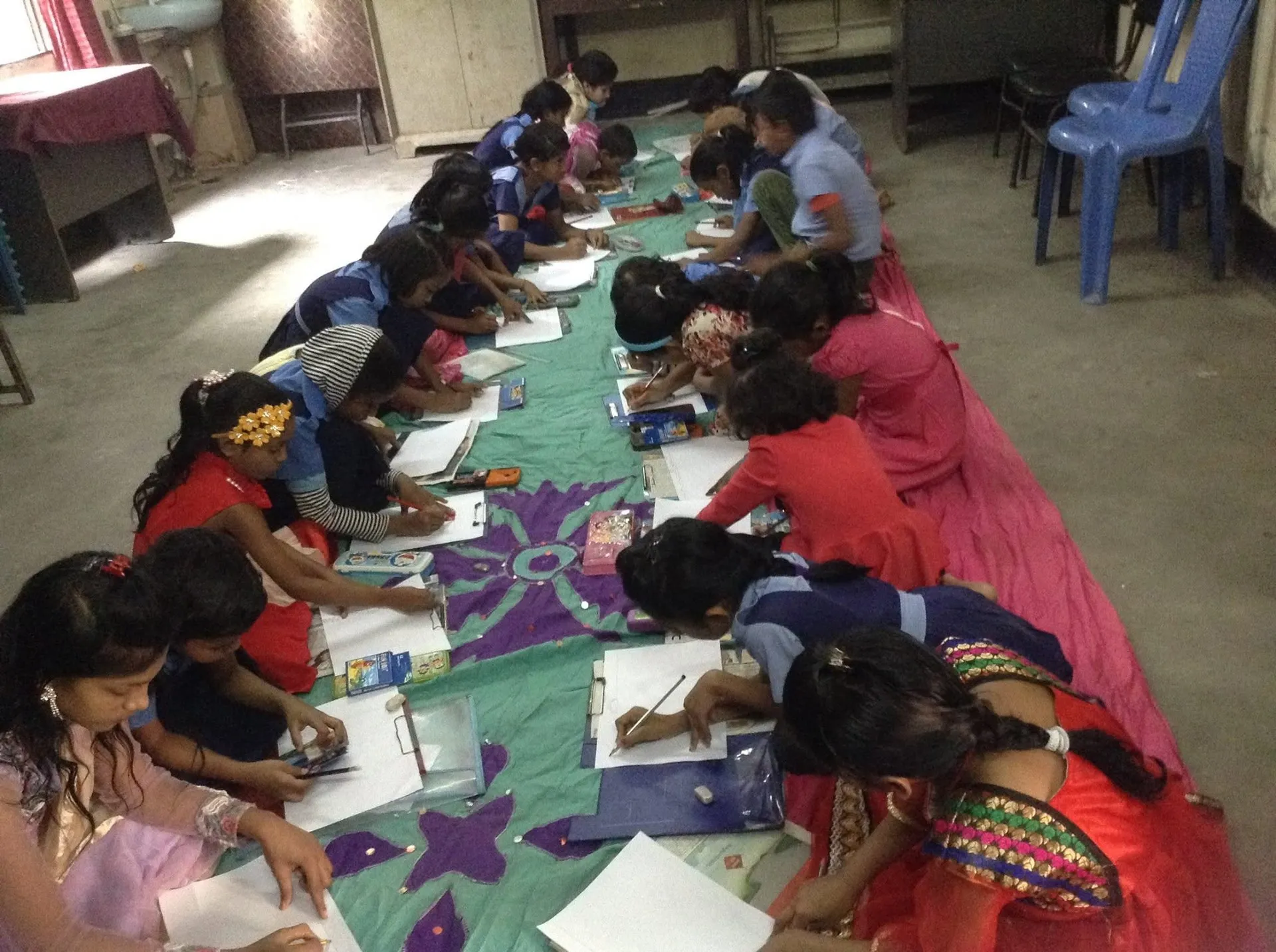 Children, some in blue school uniforms and some in festive dresses, kneel on a long mat in a classroom, drawing on sheets of paper